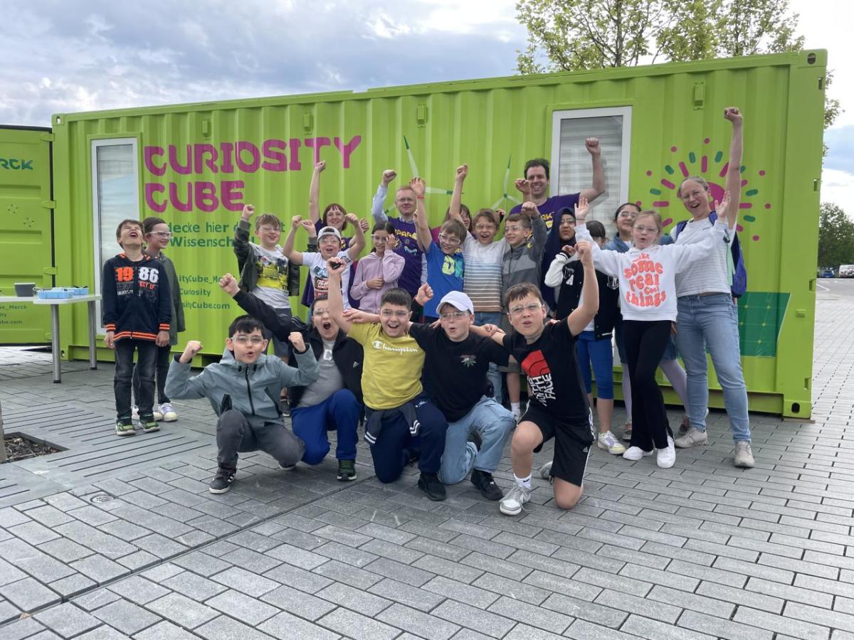 A class of students pose outdoors with MilliporeSigma employees behind the Curiosity Cube shipping container.