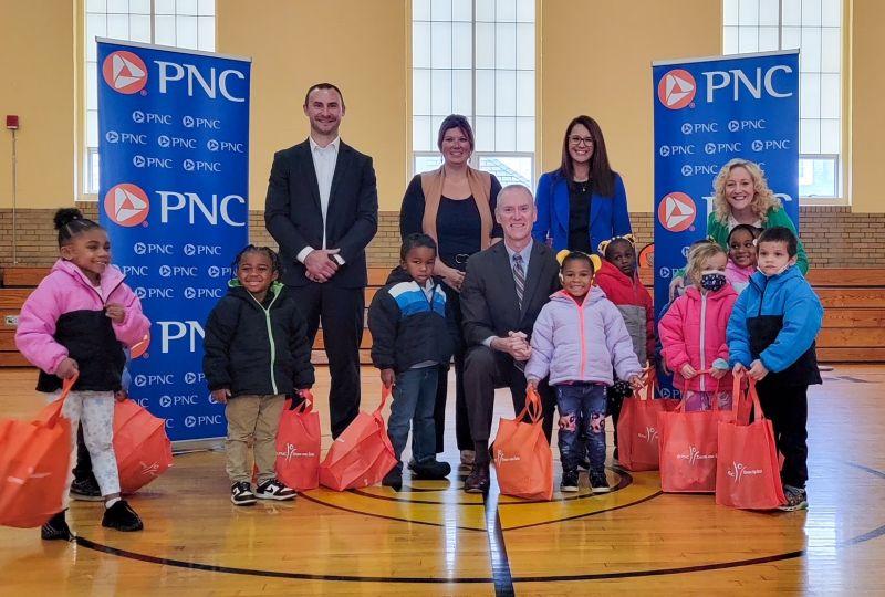 Adults and children posing in a gymnasium next to PNC banners, with orange bags