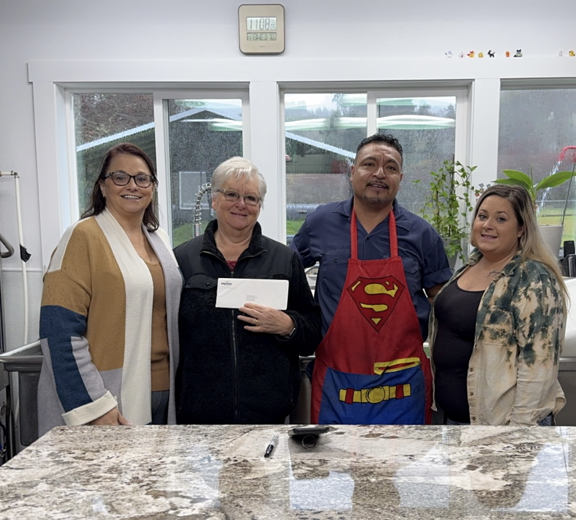 Four people standing together behind a marble table, one holding a check and one in a Superman apron