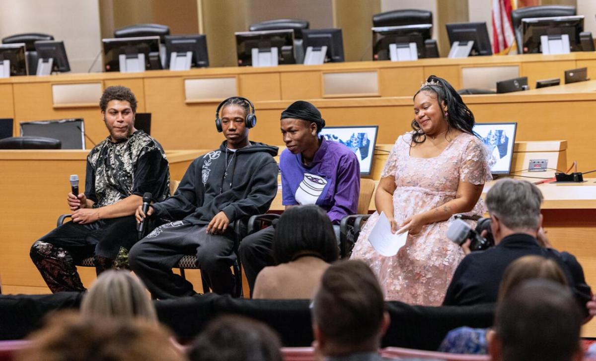 Four young people seated and facing a seated audience, two holding microphones