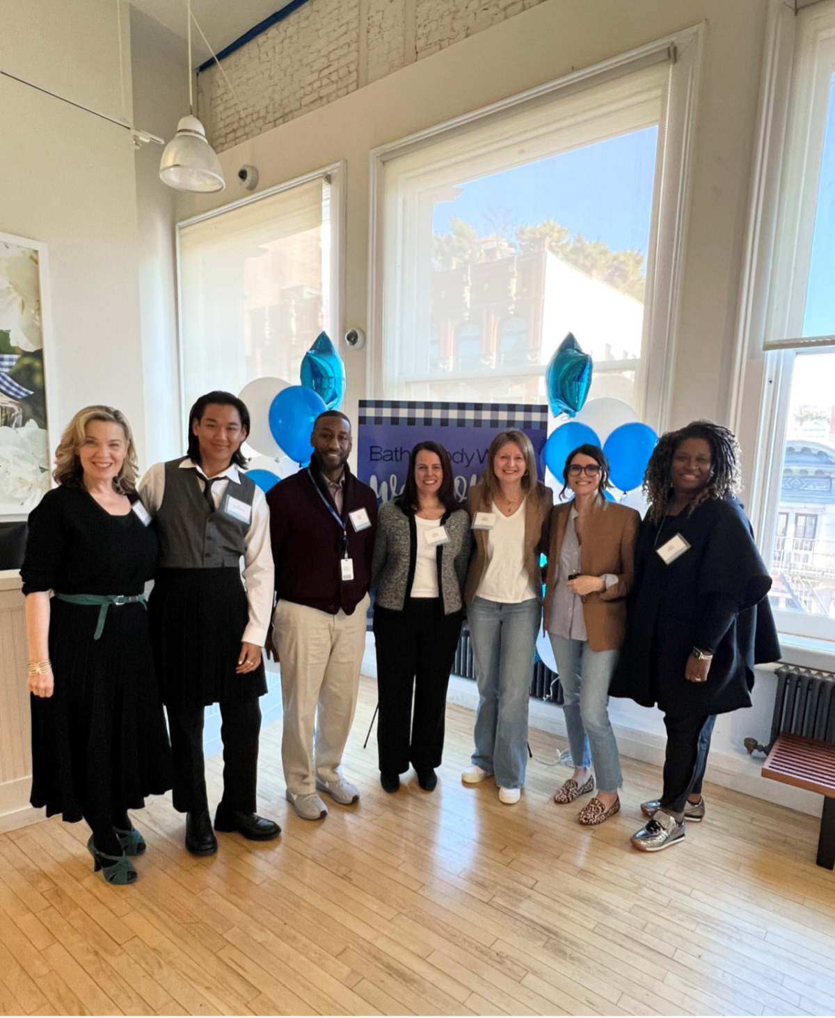 Seven Bath & Body Works employees stand together smiling at a camera in front of a sign with balloons. Sunny windows are behind the group.