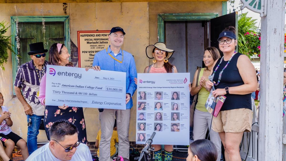 A small group, some holding a large check, and another holding a board with images of scholarship recipients.