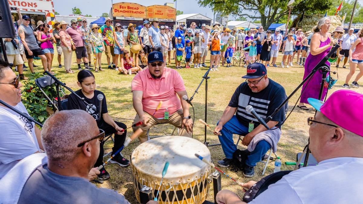 A group sitting around and beating a drum as others watch.