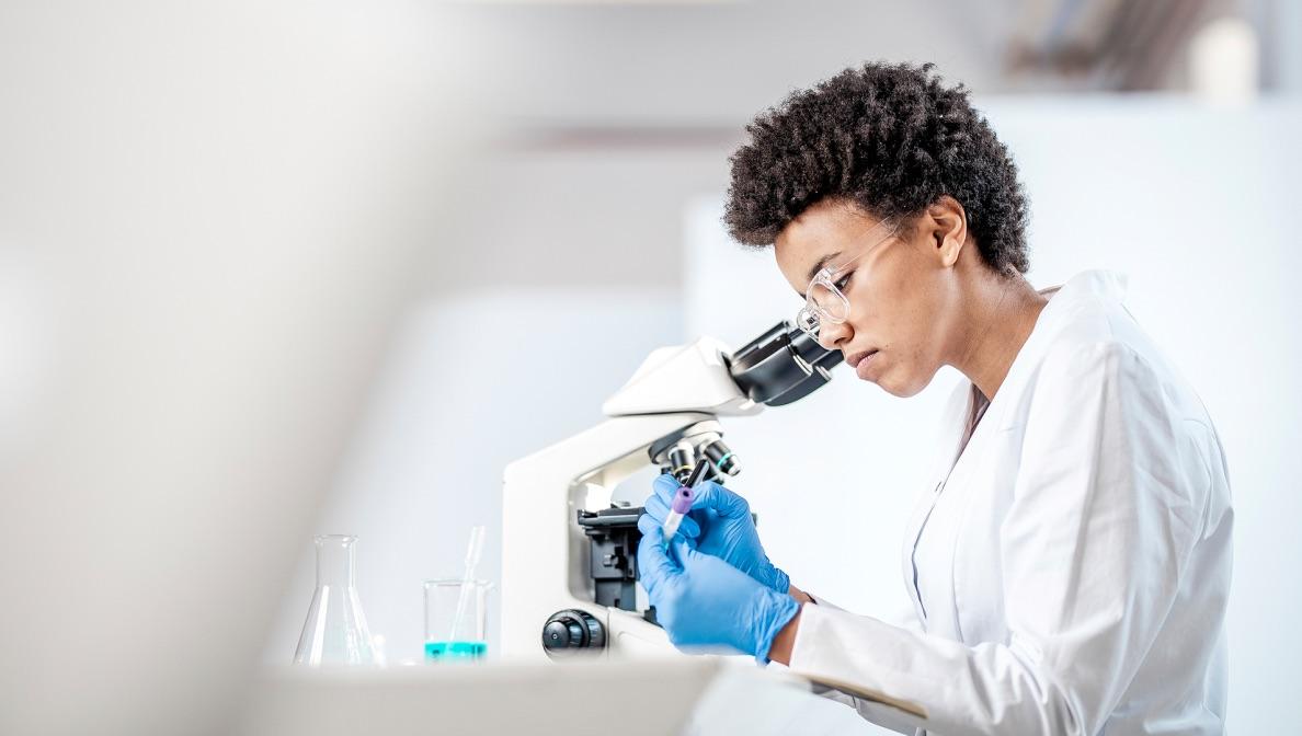 Person wearing safety goggles and a lab coat writing on a test tube next to a microscope