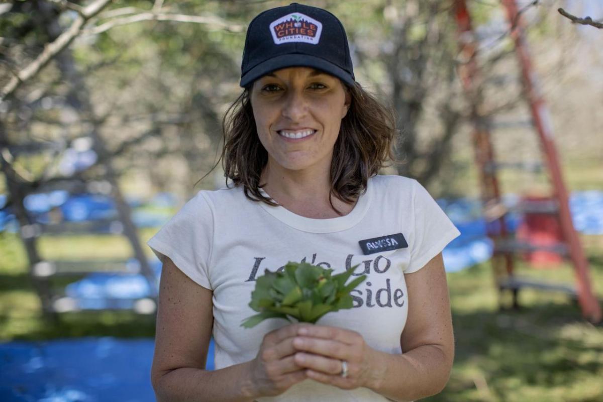 Alyssa in a white t-shirt and dark baseball cap holding a small bunch of greenery