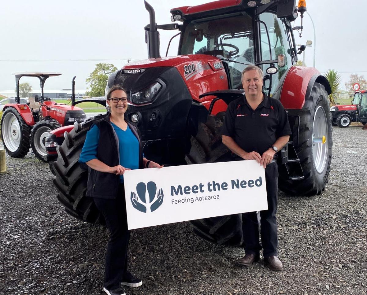 Two people stand in front of a tractor with a "Meet the Need" sign