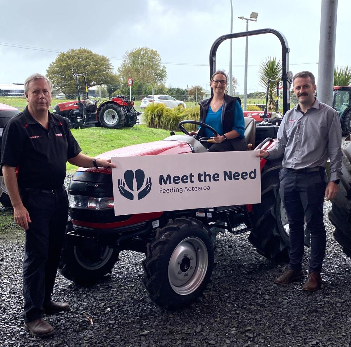 Three people stand with front of a tractor with a "Meet the Need" sign