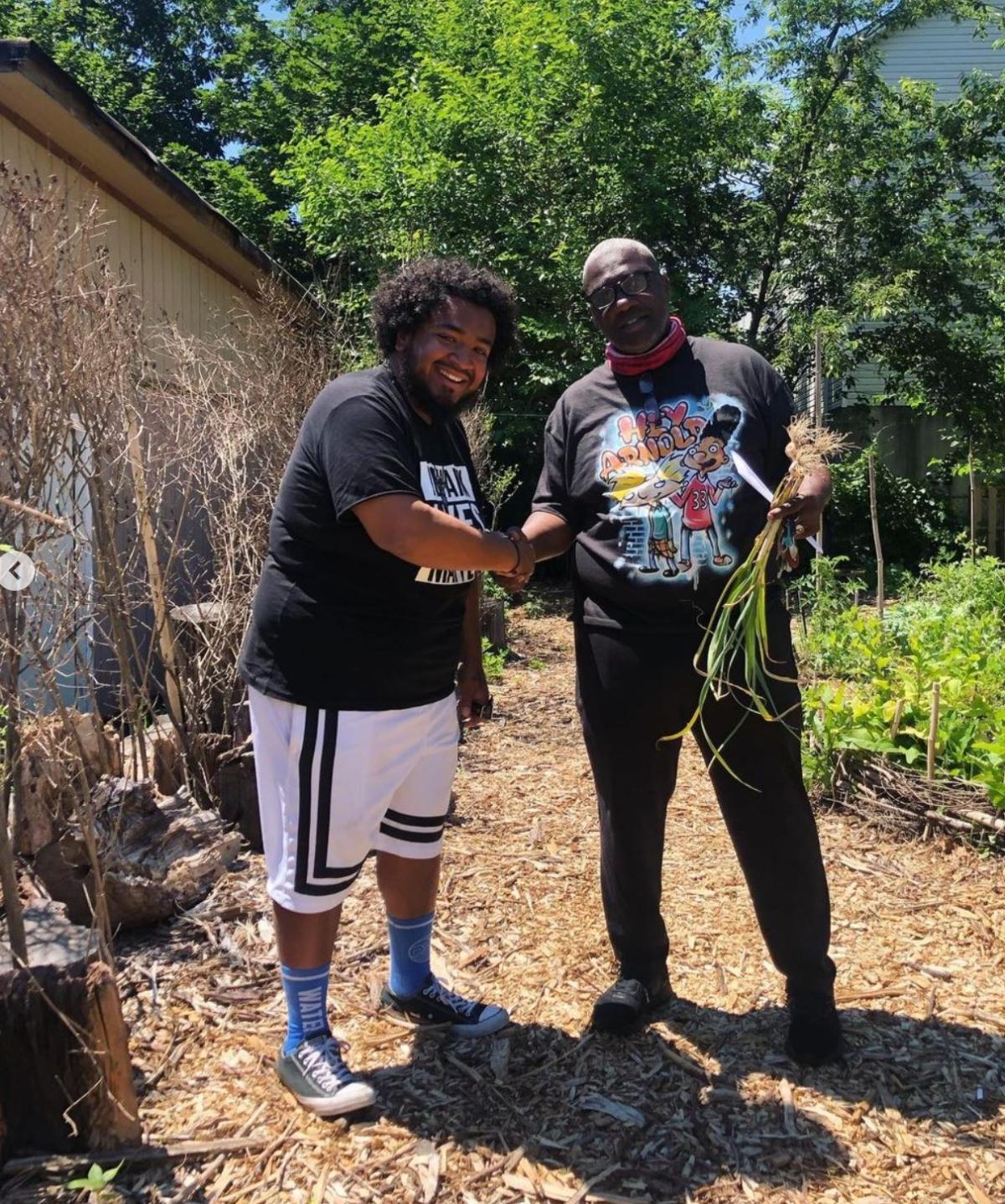 Two people shaking hands while looking at the camera. One holds a bundle of produce. They're standing on a mulch path in a vegetable garden.