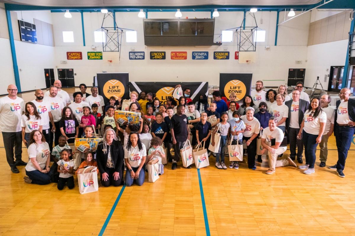 a large group of students and adults in a gym, most are wearing "Project UP" t-shirts and holding tote bags