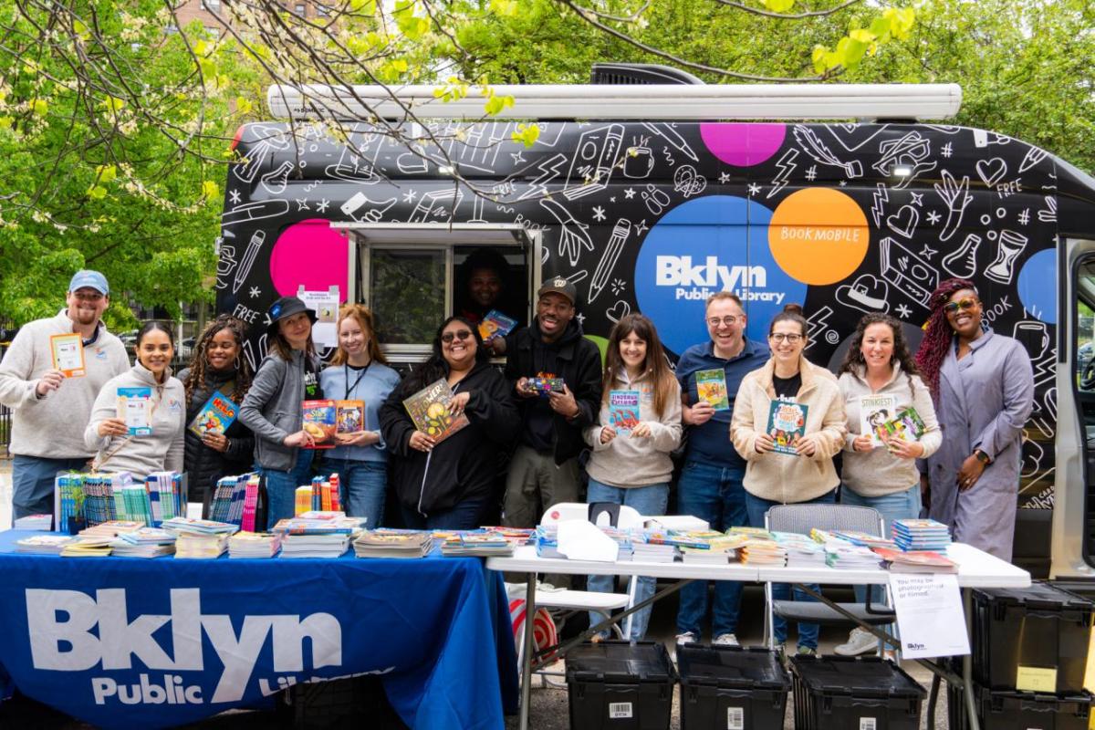 A group of people holding books, stood behind a table that has a Brooklyn Public Library banner draped over 