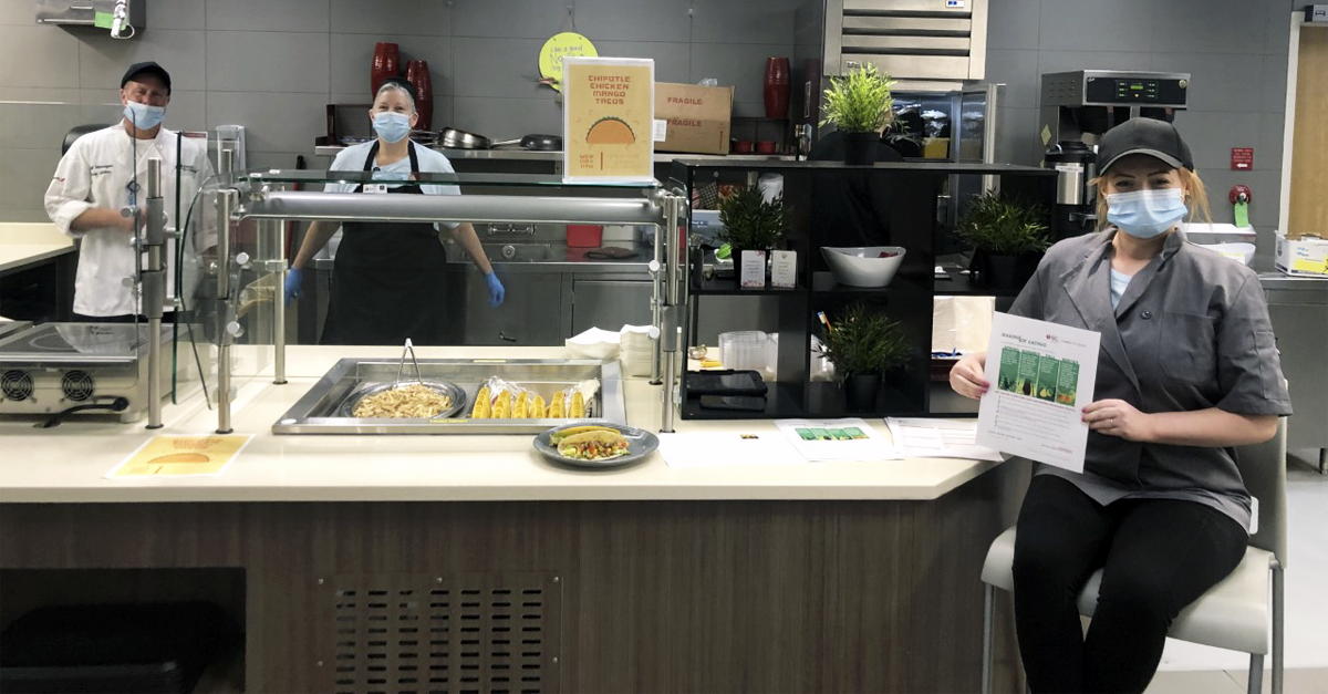 2 Blessing Hospital System masked employees stand behind a taco serving area in a cafeteria. An additional employee sits in front of the station, masked, holding up a document