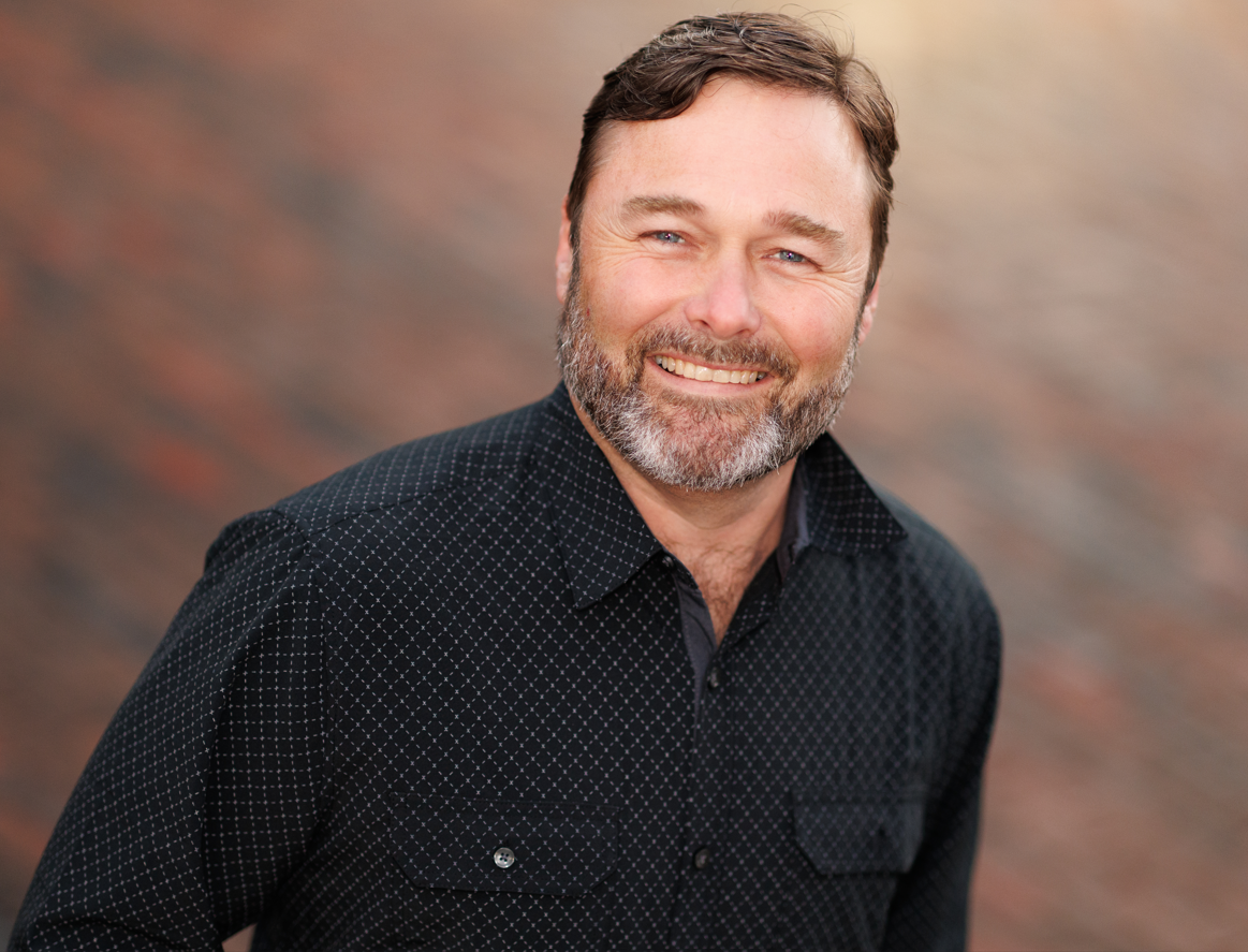Man with a beard and mustache wearing a dark patterned button-up shirt, smiling in front of a blurred brown and gray background.