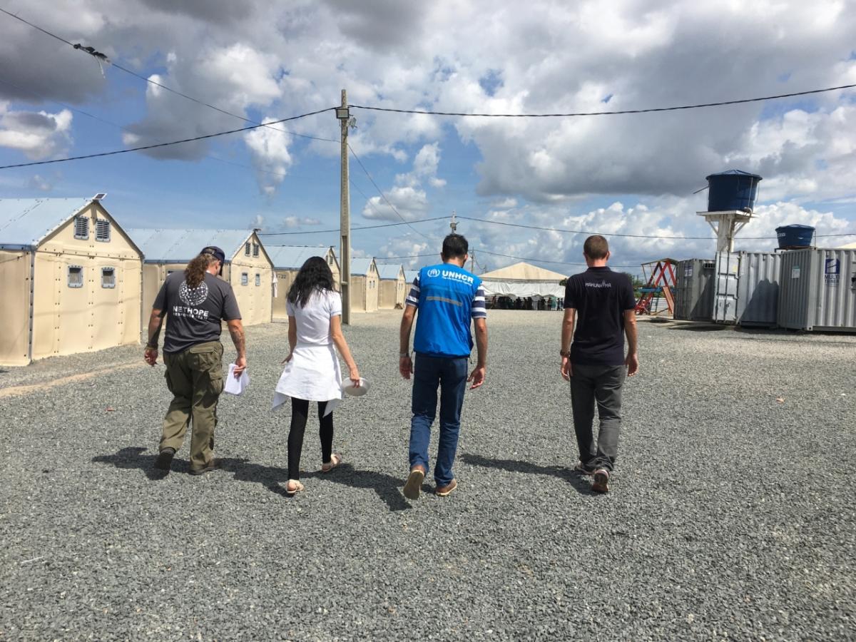 four people walking away from the camera on a gravel path. A row of Small brick buildings on their left and an open trailer on their right