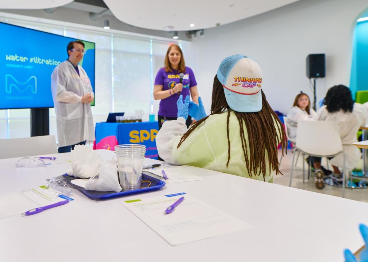 Students sit at a big table with paper and pencils while they talk with scientists at the head of a classroom.