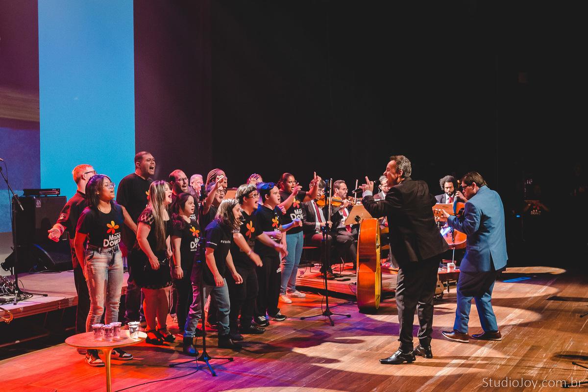 A group of students on stage with a small orchestra and conductor, all the students wearing "Mano Down" t-shirts.