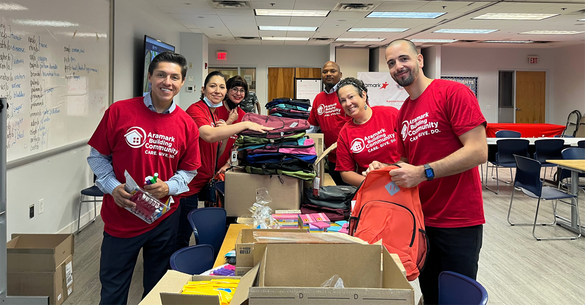 6 Aramark volunteers putting together backpacks, filling them up with assorted school supplies.