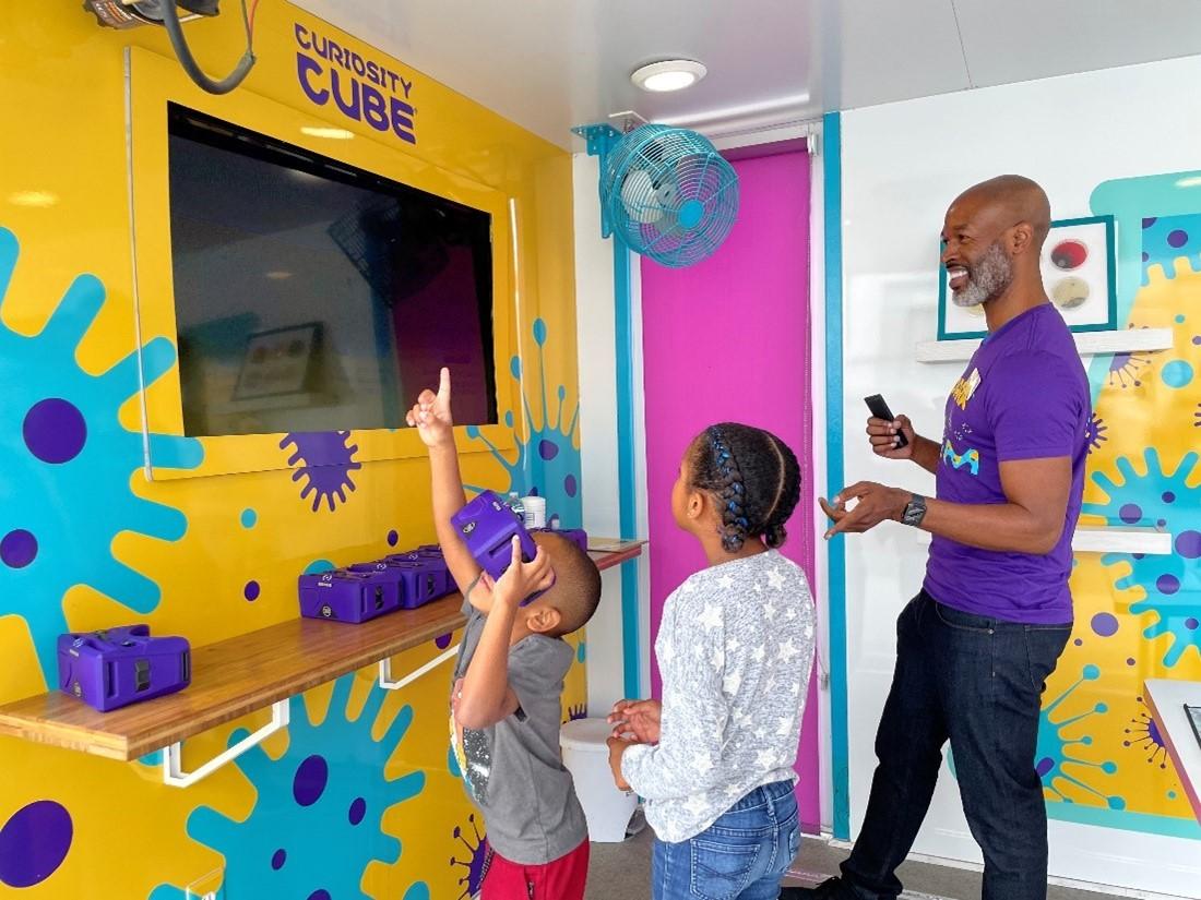 A smiling man stands next to two students who are engaging with interactive science materials. 