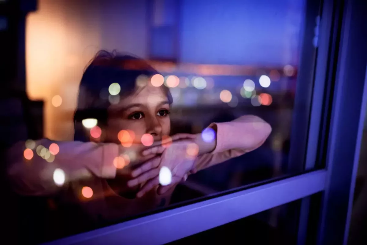 A child leant against a window with their arms folded, looking out