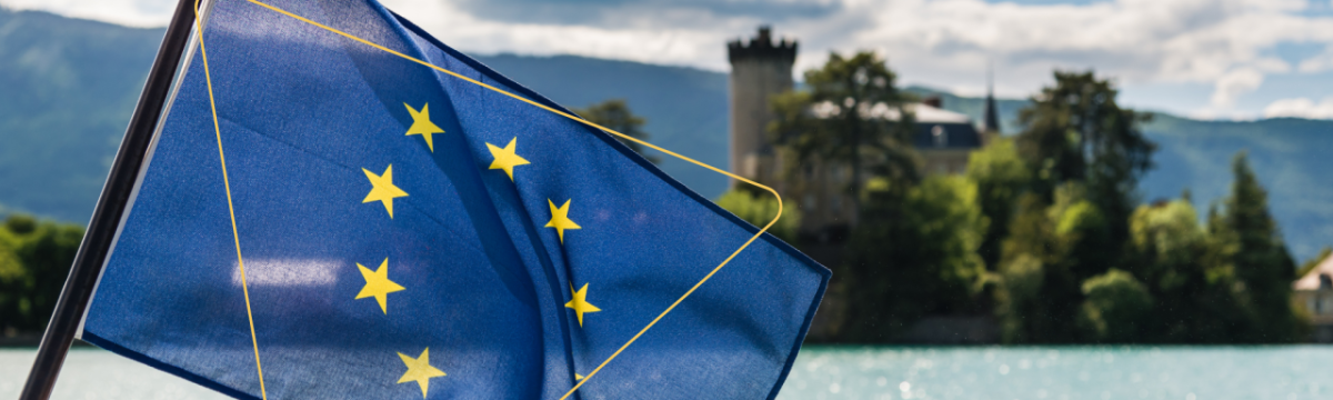 A flag and waterway, building, and scenery behind it.