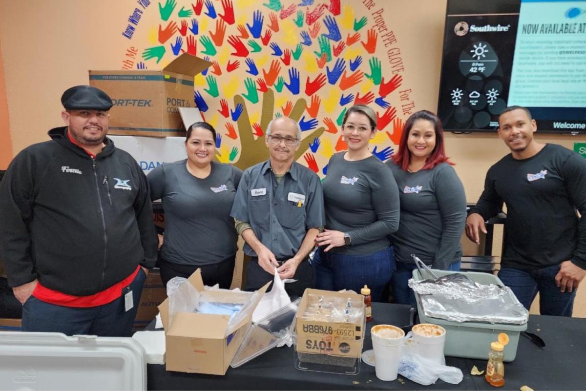 A group of employees pose for the camera, a painted mural of hands on the wall behind them and table of food and serve-ware in front of them.