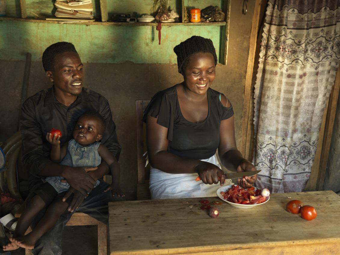 Ernest holds his youngest child in his lap while his wife cuts tomatoes.