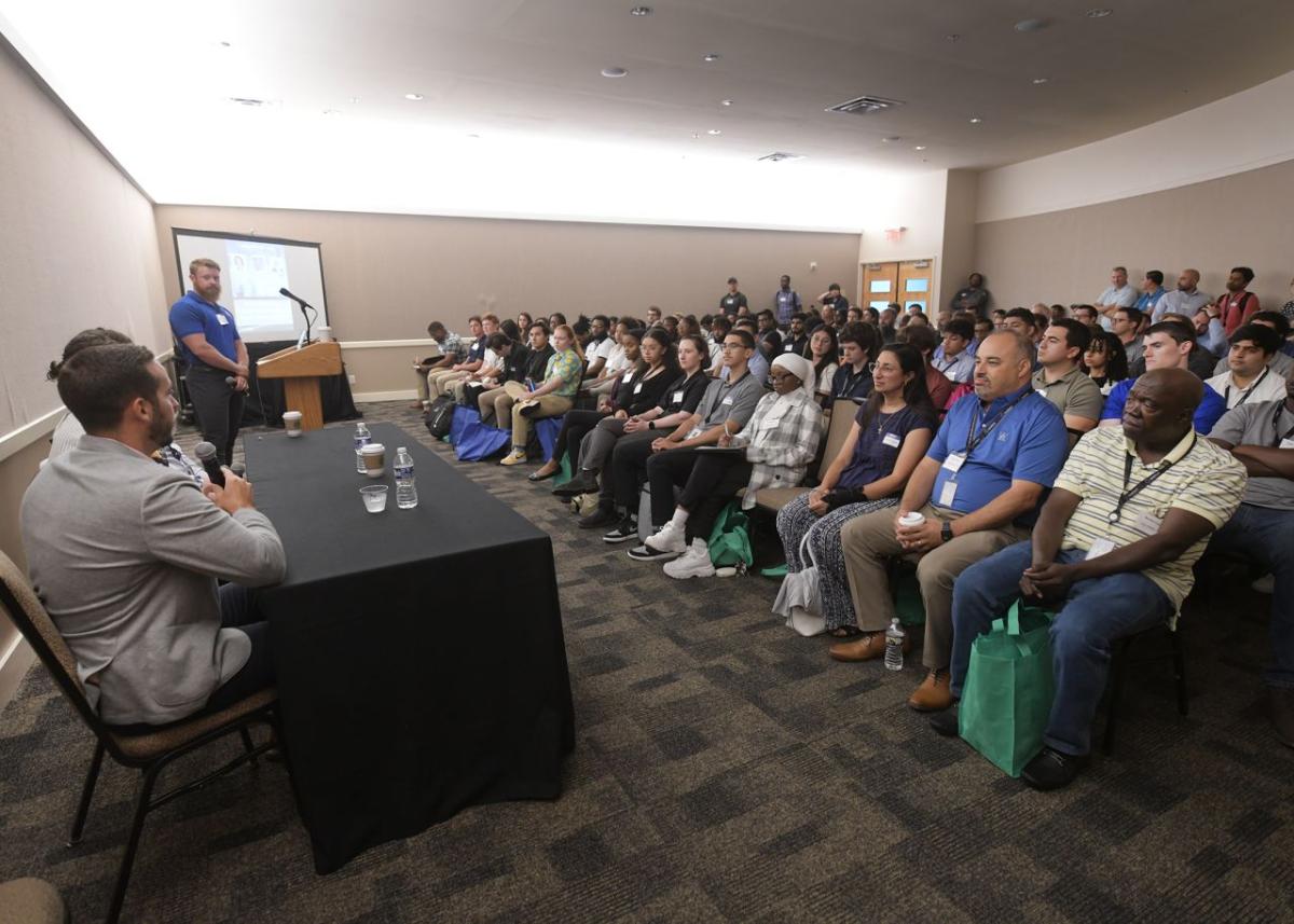 A person speaking at a podium in a room full of seated attendees. A table of other presenters at the front of the room.