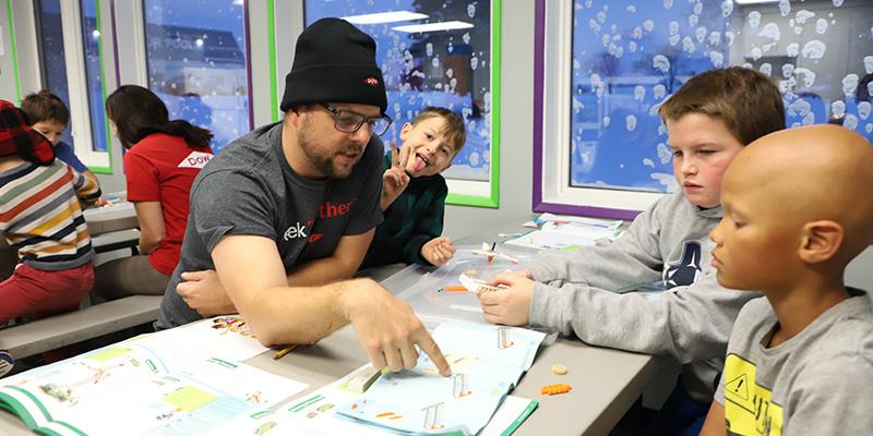 An adult pointing to instructions while three kids assemble a lego toy. Other tables doing the same.