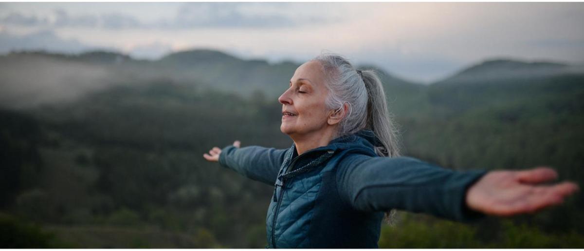 Person opens arms to sky with eyes closed with mountains behind them