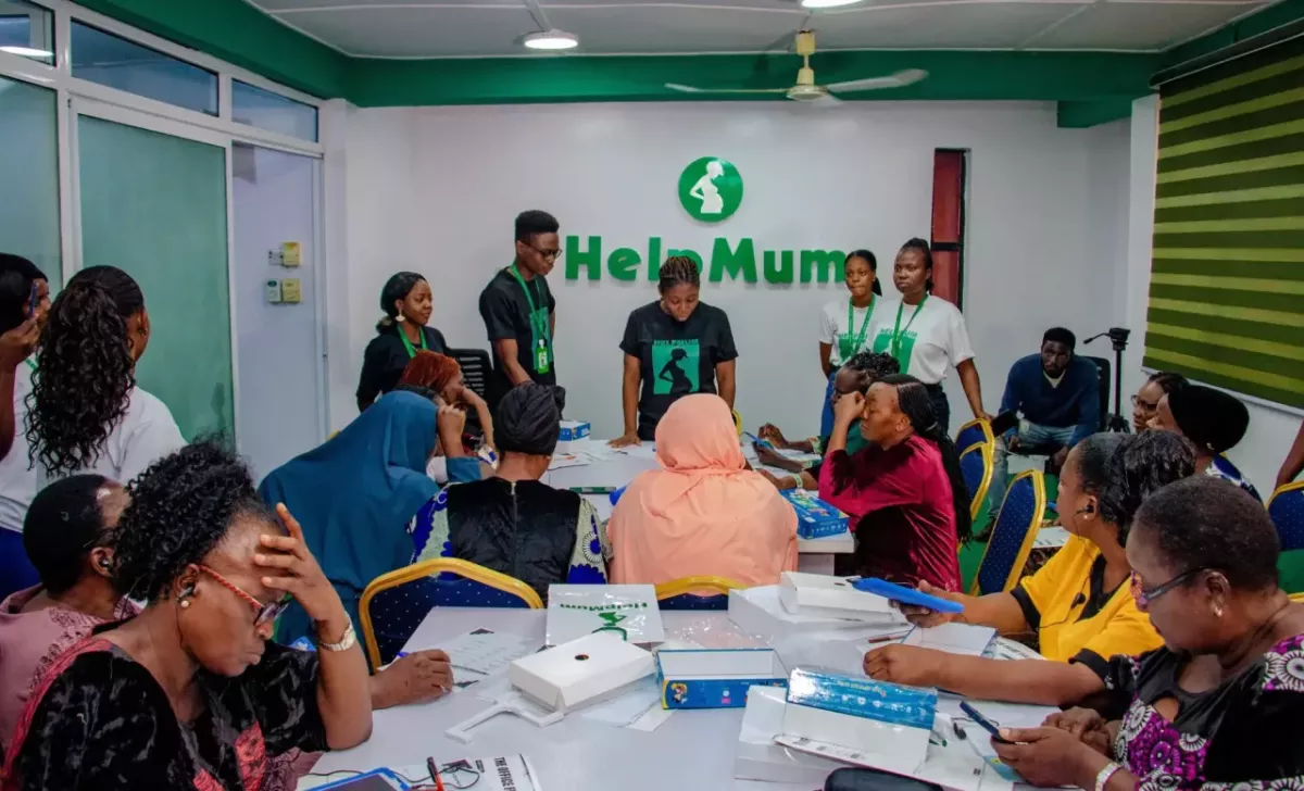 A group of people, some seated, some standing, in a conference room. A speaker at the front. "Help Mum" sign behind them.