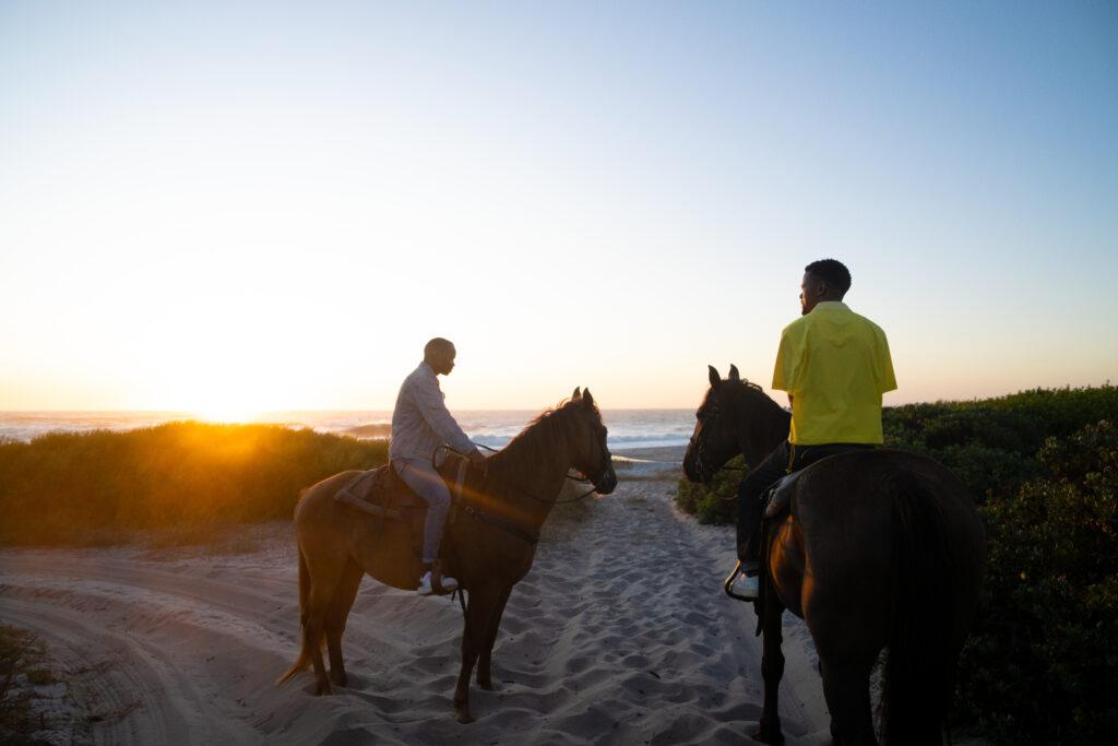 Two people horse riding on the beach, watching the sun set