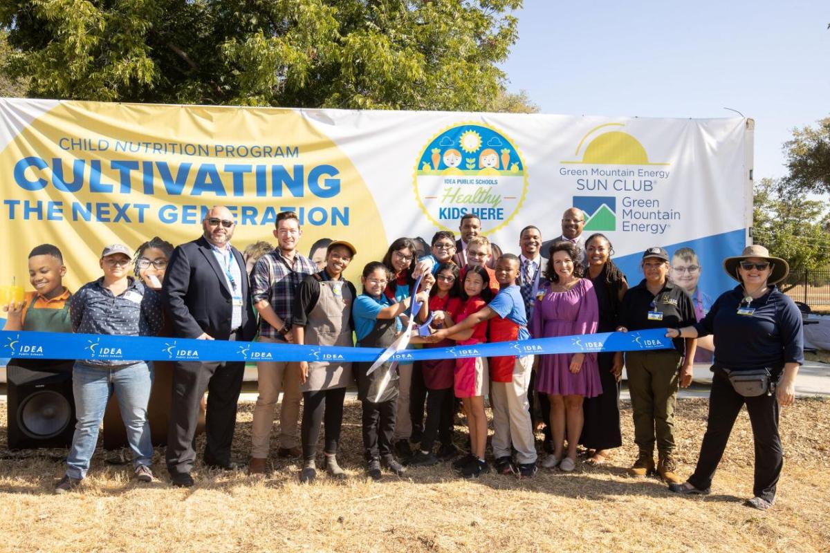 Group of people, including children, standing behind a ribbon, in front of a shipping container farm.