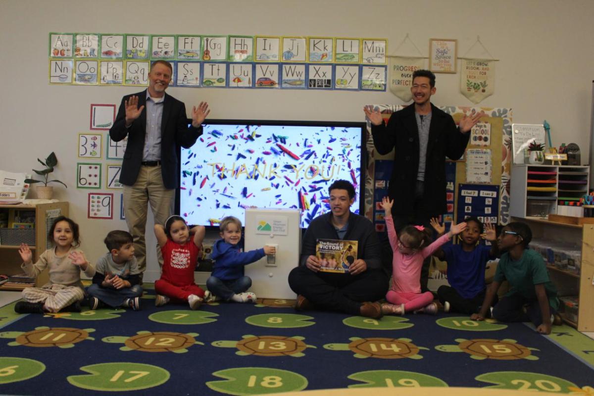 Two men stand and wave smiling, while children sit around an oversized prop light switch cheering