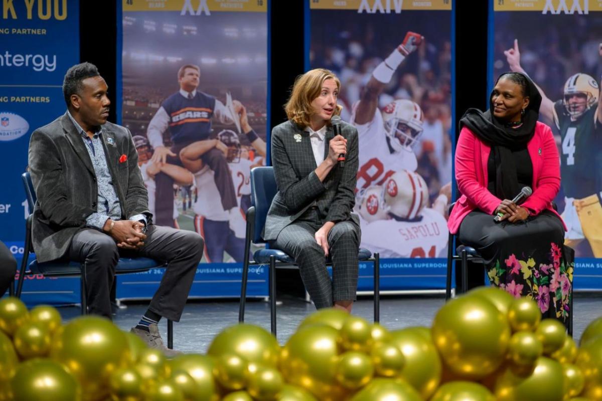 Three speakers seated on a stage, one speaking into a microphone.