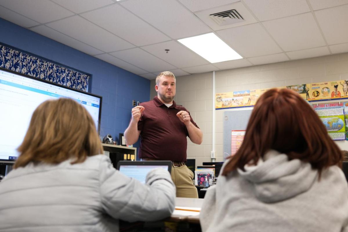 Classroom with students sat at a desk while a teacher is stood in front of the class 