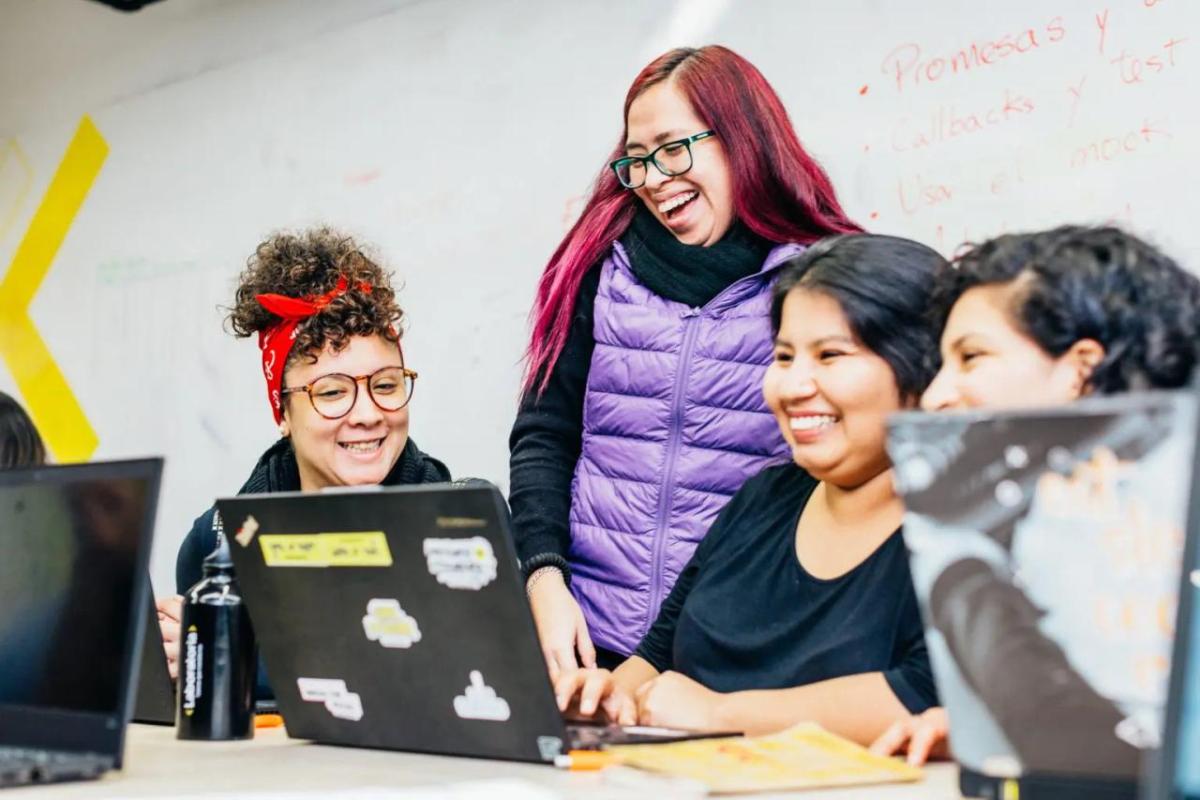 Three smiling people seated, looking at a laptop, a fourth behind them. A whiteboard with writing on it in the background.