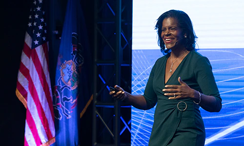 Lynnette speaking with hand gestures, two flags to the left and a large digital screen behind.