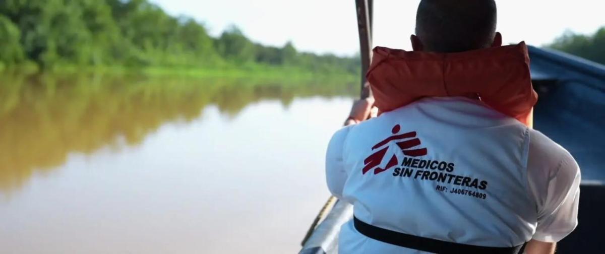 A person wearing a life vest, on a boat in a body of brown water. Trees line the shore.