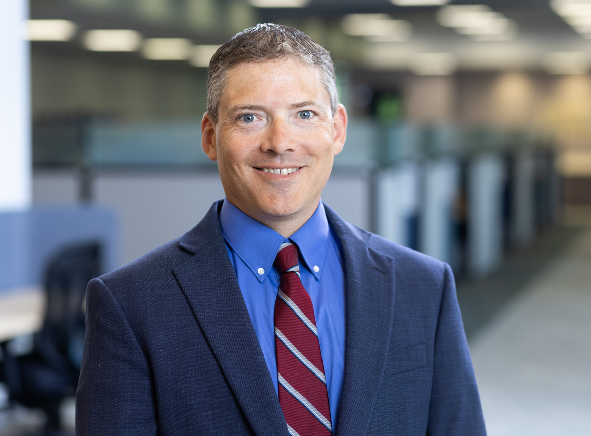Man wearing a navy suit, blue shirt, and red striped tie, smiling in an office setting with cubicles in the background.