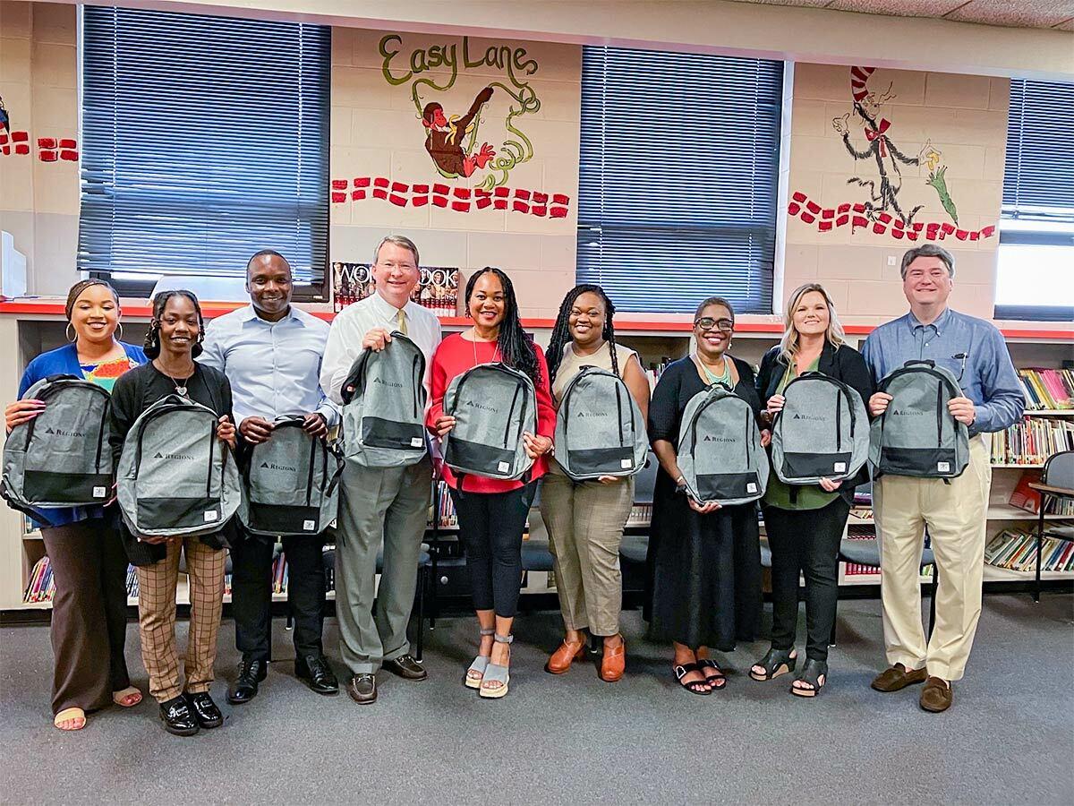 A line of people in a school setting, each holding a grey backpack.