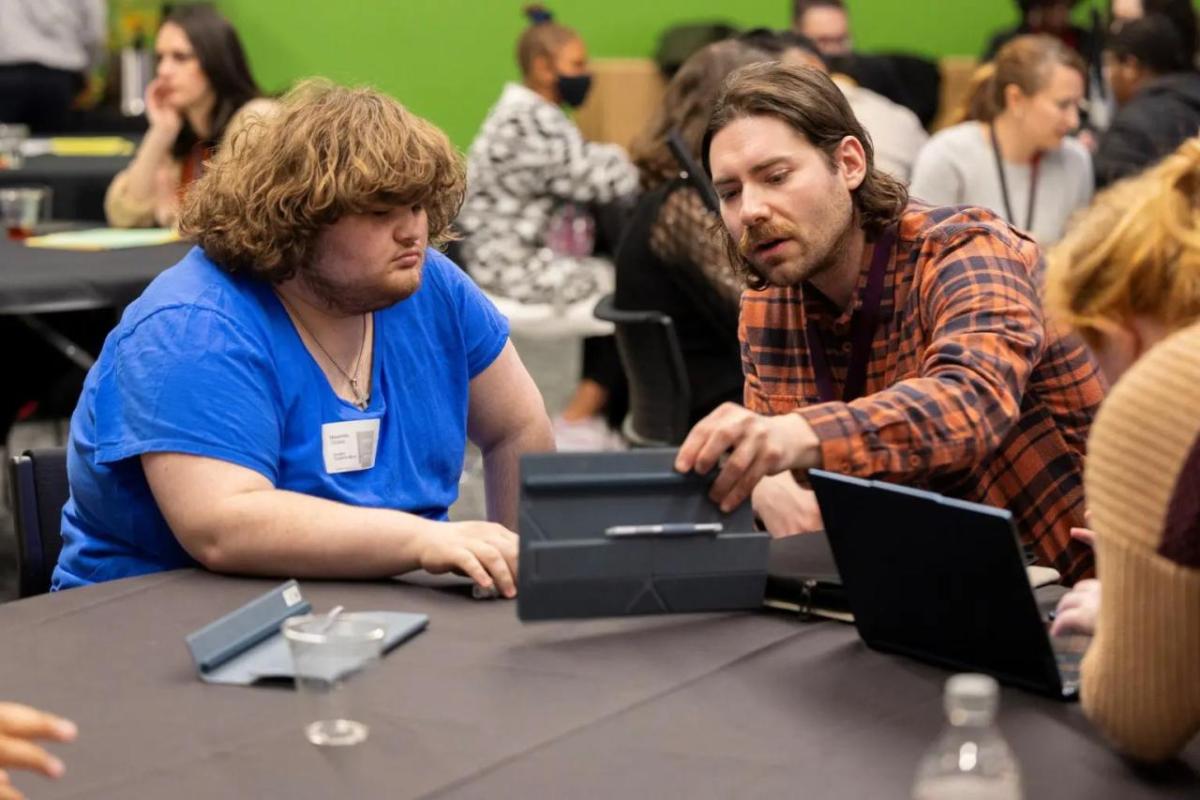 Two people looking at an open laptop in a room of others seated at different tables.