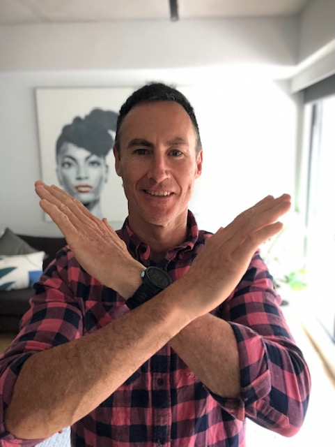 Man in a plaid shirt, standing in front of a picture of a woman, crossing his arms