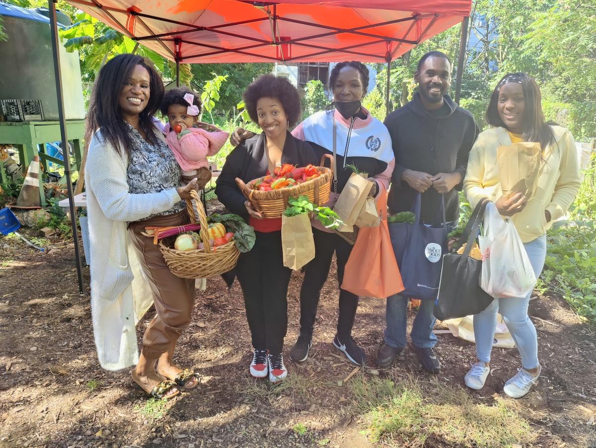 A group of people including a young child being held, each carry bags and baskets of fresh produce. Standing under a red pop-up awning outside.