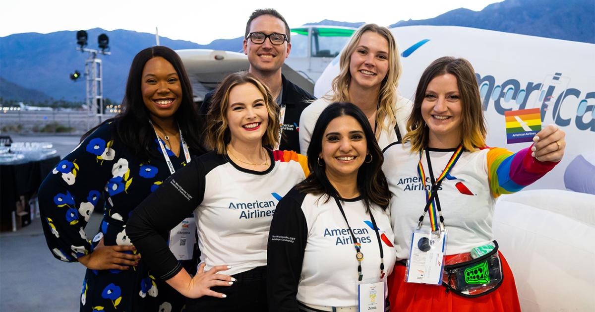 group of six people standing with planes in the background. One holds a small rainbow flag.