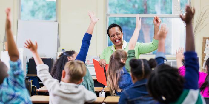 Female teacher in front of her classroom of children. The children have their hands raised.