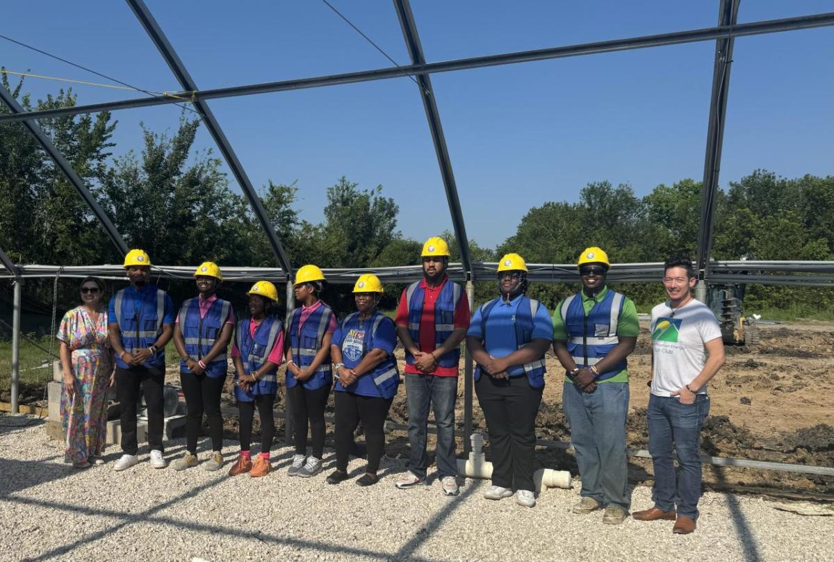 Group of people in greenhouse structure, wearing blue construction vests and yellow hard hats