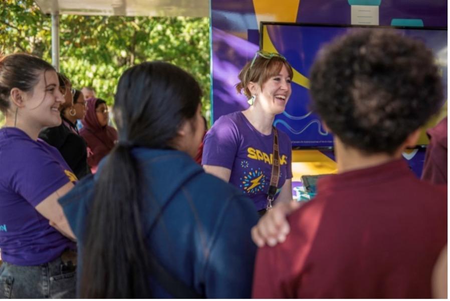 A woman in a MilliporeSigma Spark T-Shirt smiles alongside students. 