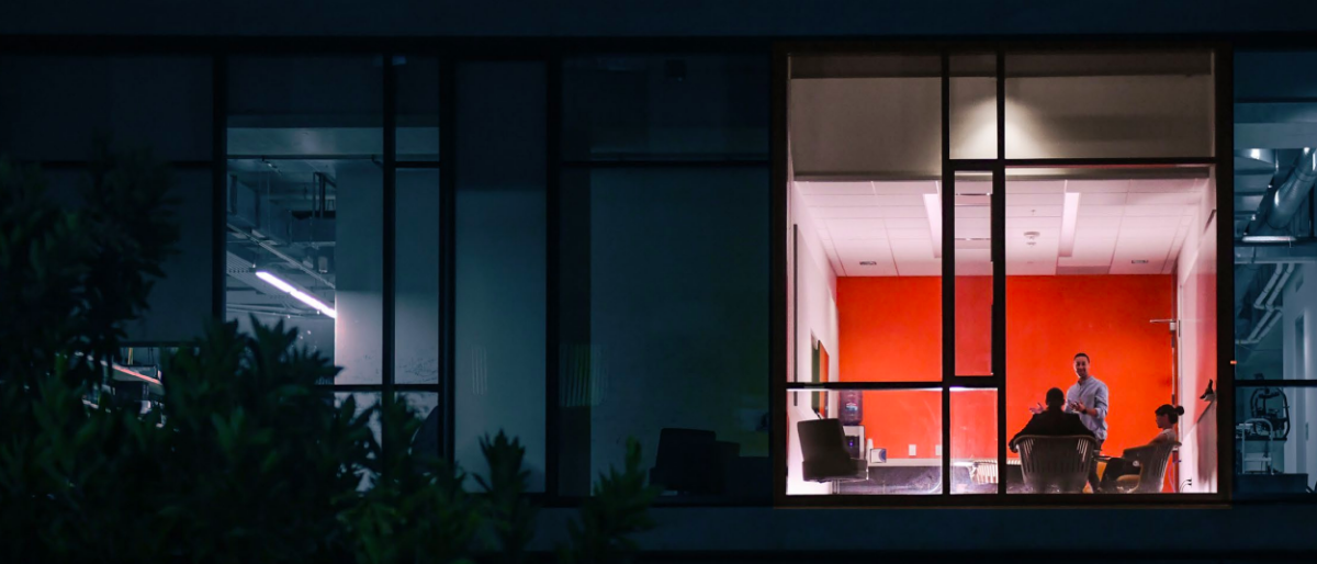 Looking in from the outside at two people talking in a lit office space, other spaces in the building are dark.