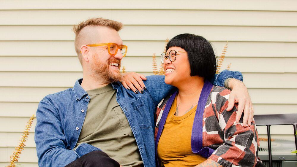 Two seated people smiling at each other with an arm around each other.