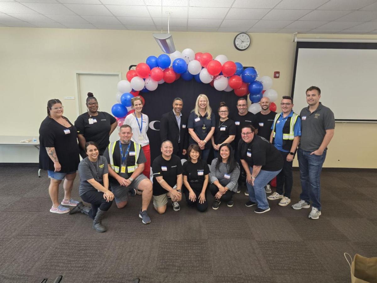 A group of men and women posing for a photo in front of a black backdrop with an arch of red, white, and blue balloons overhead.