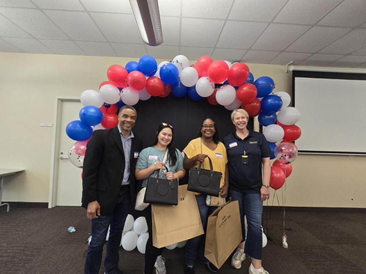 A man and three woman posing for a photo in front of a black backdrop with an arch of red, white, and blue balloons overhead.